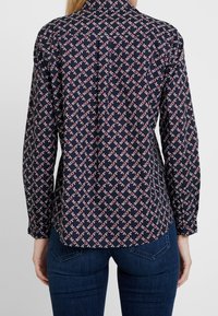 Blusa a fantasia navy con un design geometrico in rosso e bianco, maniche lunghe, colletto a punta e orlo curvo con una texture liscia.