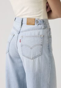 Personne portant un jean Levi's bleu clair taille haute avec des poches arrière visibles, un patch en cuir de la marque et un haut blanc rentré.