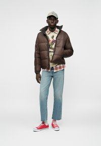 Brązowa kurtka puchowa, wzorzysty sweter, koszula w kratę, jasnoniebieskie dżinsy i czerwone sneakersy. Na głowie szara czapka; casualowy strój.