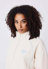 Felpa zip con crema e dettagli blu chiaro. Presenta un collo alto e un logo ricamato sul petto. Texture morbida.