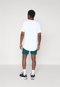 T-shirt blanc à manches courtes avec un ourlet arrondi, associé à un short de sport vert foncé. Modèle portant des baskets noires avec des accents blancs.