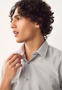 Jeune homme aux cheveux bouclés ajustant le col d'une chemise boutonnée à motifs, regardant vers la gauche sur un fond uni.