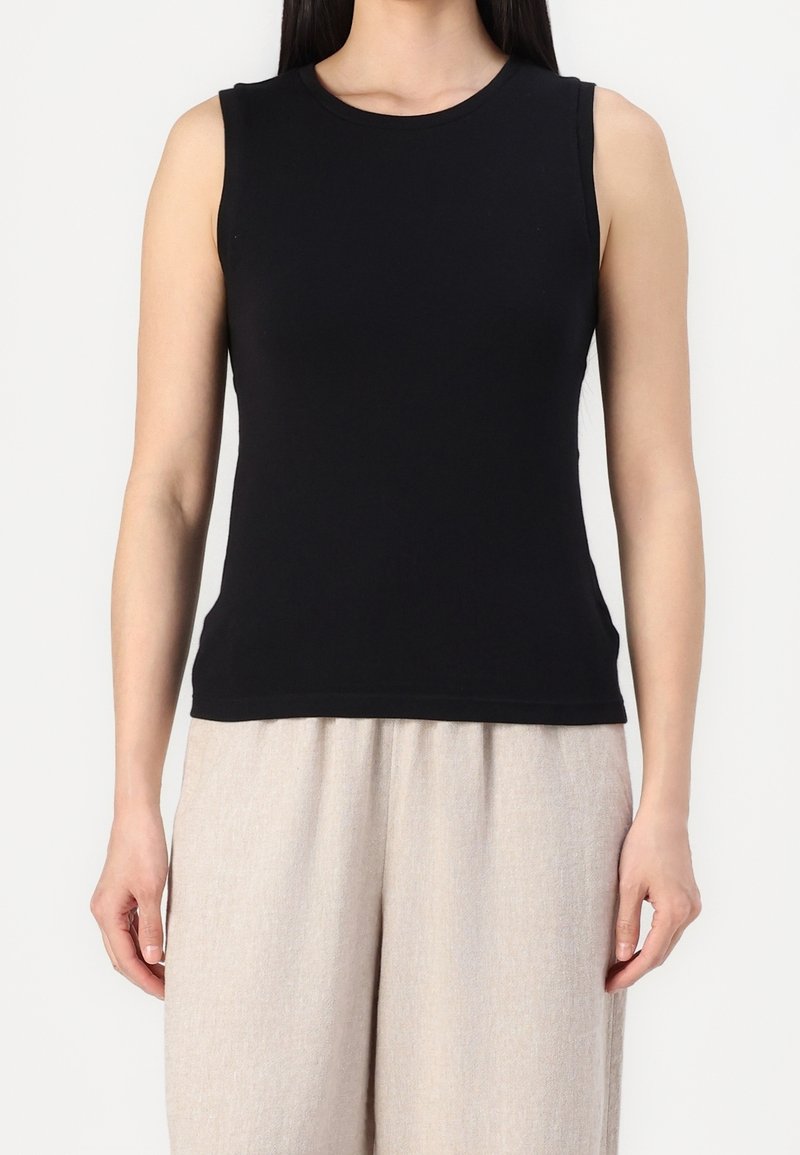 Mujer con una blusa negra ajustada sin mangas combinada con pantalones sueltos beige claros, frente a un fondo blanco liso.