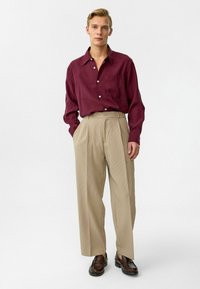 Marsala button-down overhemd, beige geplooide broek, soepele textuur, losse pasvorm, gecombineerd met bruin leren instappers op een effen lichte achtergrond.