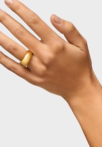 Złoty metalowy pierścień o gładkim, zaokrąglonym wzorze, noszony na ręce z lekko wypielęgnowanymi paznokciami.