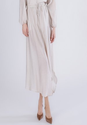 Kobieta w długiej, jasnobeżowej sukni z marszczonym stanem i długimi rękawami, w parze z brązowymi, spiczastymi butami na wysokim obcasie.