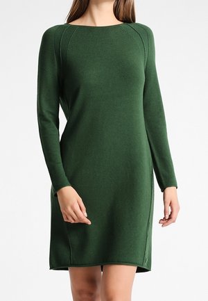 Frau trägt ein dunkelgrünes, gestricktes Kleid mit langen Ärmeln und knielang vor einem schlichten weißen Hintergrund.