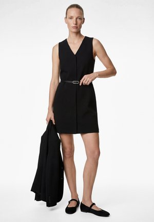 Kobieta w czarnej, bezrękawnej sukience z dekoltem w kształcie litery V i paskiem, nosząca czarne baleriny, trzymająca czarną marynarkę w lewej ręce, stojąca na białym tle.