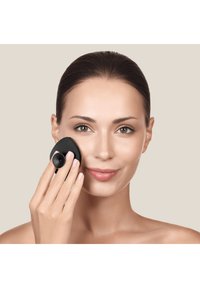 Strumento di pulizia del viso nero con un design arrotondato, bordi textured e una funzione di aspirazione, tenuto contro la guancia di una donna. Sfondo di pelle liscia.