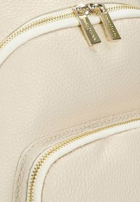 Béžový kožený batoh s texturovaným povrchem, vybavený zlatým kováním a dvojitými zipy. Zipy jsou lemovány bílou látkou.