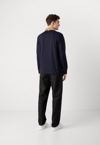 Camicia a maniche lunghe blu marina con colletto beige, abbinata a pantaloni scuri e sneakers bianche. Presenta un design semplice senza pattern visibili.
