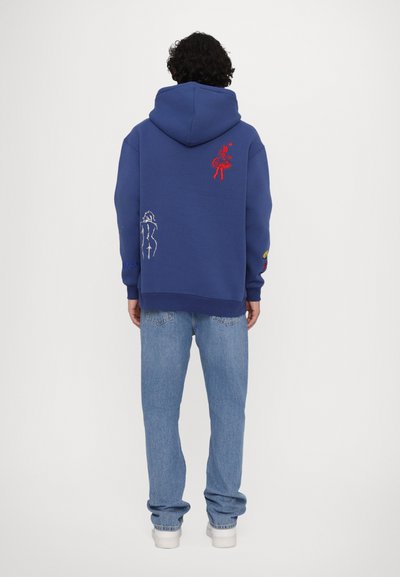 Felpa blu con design ricamati sul retro, che presenta una figura rossa e una figura bianca. Abbinata a jeans azzurri e scarpe bianche.