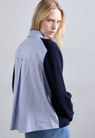 Camicia a righe blu con maniche navy, caratterizzata da un design con pieghe sul retro e un colletto classico. Tessuto morbido con texture a contrasto.