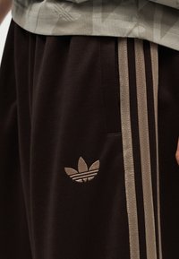 Κοντινό πλάνο σκούρου καφέ παντελονιών Adidas με μπεζ ρίγες και λογότυπο, φορεμένα με μπεζ τοπ.