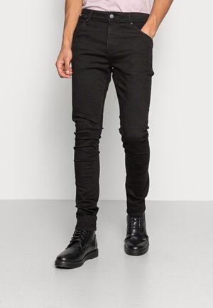 Mann trägt eng anliegende schwarze Jeans und schwarze Schnürstiefel, steht mit einer Hand in der Tasche.