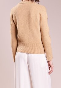 Helles beige Strickpullover mit geripptem Saum, strukturiertem Stoff und Ellenbogen-Ausschnitten. Kombiniert mit weißen weit geschnittenen Hosen.