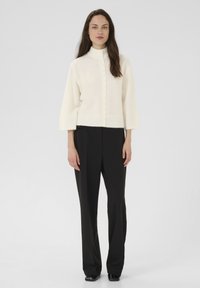 Nő áll szemben, fehér, gombos pulóverben, széles ujjakkal, és fekete, bő szárú nadrágban, egy sima háttér előtt.