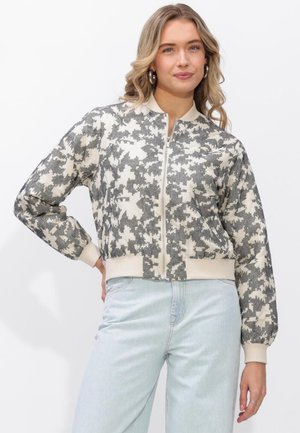 Vrouw draagt een grijs-wit bloemenjack met rits en lichte blauwe spijkerbroek, staand met één hand op haar heup tegen een witte achtergrond.