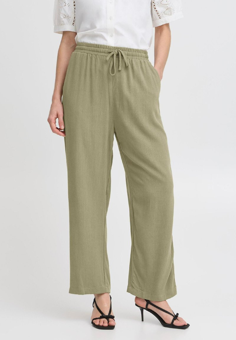 Pantalons larges vert olive clair avec une taille élastique et un cordon de serrage, en tissu doux. Associés à des escarpins à talons hauts noirs avec des lanières.