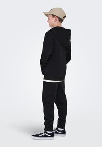 Sort hættetrøje og sweatpants, begge med ribbede manchetter, iført en beige kasket og sorte sneakers. Enkel design, stoftekstur ikke synlig.