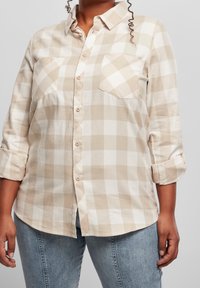 Persona che indossa una camicia a quadri beige e bianca con maniche arrotolate e jeans azzurri chiari su uno sfondo neutro.