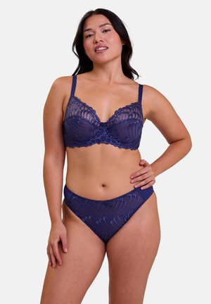 Femme portant un soutien-gorge en dentelle bleu marine et un slip taille haute assorti, debout avec une main sur la hanche sur un fond uni.