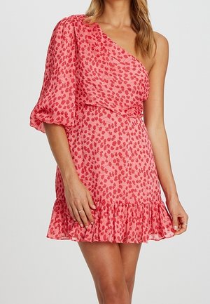 Kobieta w różowej sukience na jedno ramię z czerwonymi kwiatowymi nadrukami, bufiastym rękawem i falbaną na dole, stojąca na jednolitym tle.