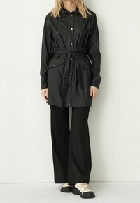 Zwarte waterdichte parka met een capuchon met trekkoord, drukknoopsluiting, twee voorzakken en een ceintuur in de taille. Geroeid met bijpassende zwarte broeken en beige sneakers.