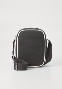 Black fabric crossbody bag with white trim, embossed Lacoste logo, rectangular shape, adjustable strap, and zip closure.