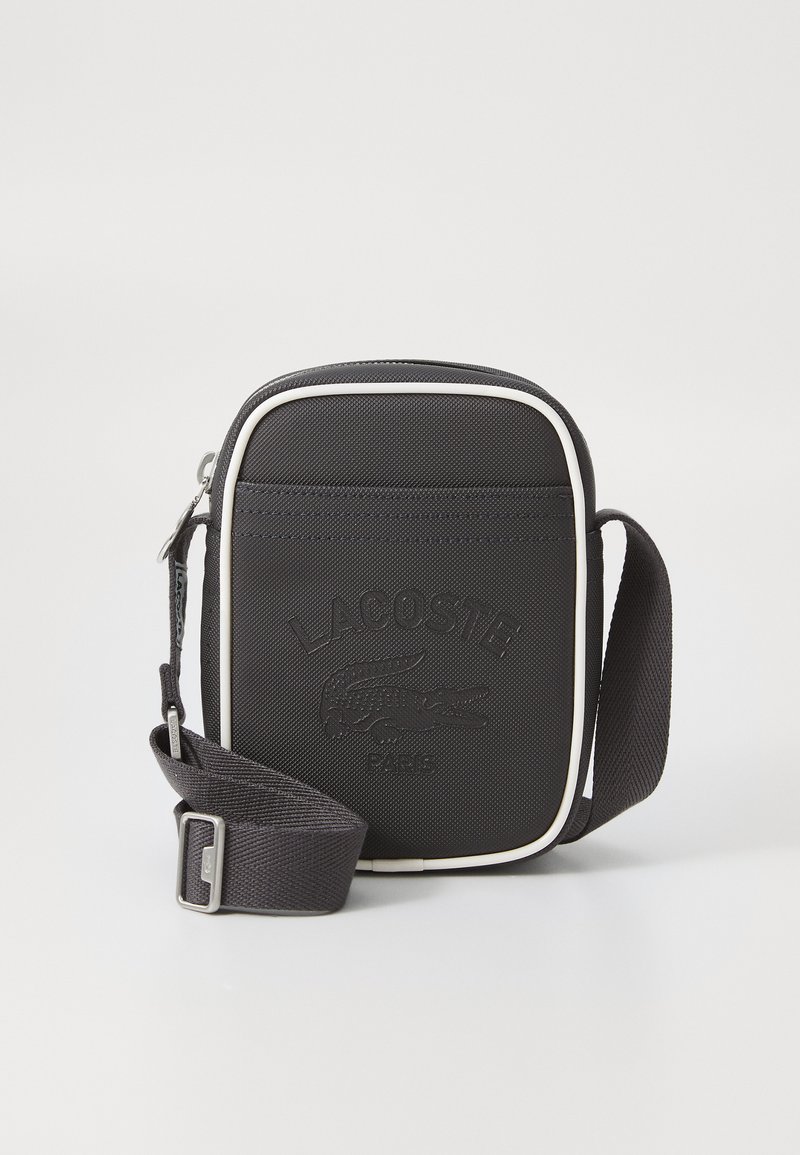 Black fabric crossbody bag with white trim, embossed Lacoste logo, rectangular shape, adjustable strap, and zip closure.