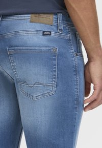 Jeans de denim azul con un acabado desgastado. Presenta un bolsillo trasero con un detalle de costura en zigzag y una etiqueta de cuero en la cinturilla.