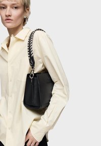 Młoda osoba w kremowej koszuli z guziczkami, nosząca perłowe kolczyki i trzymająca czarną torebkę na ramię z paskiem w czarno-białe paski.