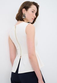 Weißes Peplum-Top mit goldener Rückenzipper, ärmellosem Design und strukturiertem Finish. Mit einem Schlitz auf der Rückseite für zusätzliche Details.