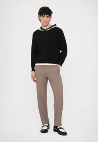 Fekete kötött pulóver csíkos gallérral, bézs nadrággal és fekete-fehér cipővel párosítva. A modell fehér háttér előtt áll.