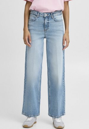 Persoon die lichtblauwe, hoge taille, wijde jeans draagt, gecombineerd met witte sneakers en een ingestopt roze T-shirt.