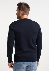 Sötétkék kötött pulóver hosszú ujjakkal, bordázott nyakkal, mandzsettával és aljjal. Világoskék farmernadrággal viselve, sima textúrával.