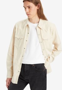 Camisa de mezclilla beige con botones a presión y dos bolsillos en el pecho, llevada sobre una camiseta blanca, combinada con jeans negros.