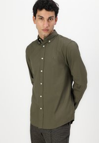 Camicia a maniche lunghe verde oliva con colletto button-down, bottoni bianchi e un piccolo logo sul petto. Tessuto dalla texture liscia.