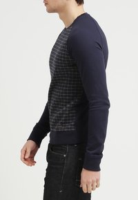 Hombre vestido con sudadera navy con patrón de cuadrícula geométrica en el frontal, mangas lisas y pantalones vaqueros oscuros, de pie de perfil contra un fondo blanco.
