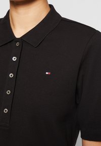 Schwarzes Poloshirt aus strukturiertem Material, mit einem klassischen Kragen, drei Knöpfen und einem kleinen gestickten Logo mit rot, weiß und blauem Akzent.