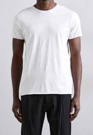 Man die een effen witte korte mouw t-shirt en zwarte krijtstreep broek draagt, staand tegen een lichtgrijze achtergrond.