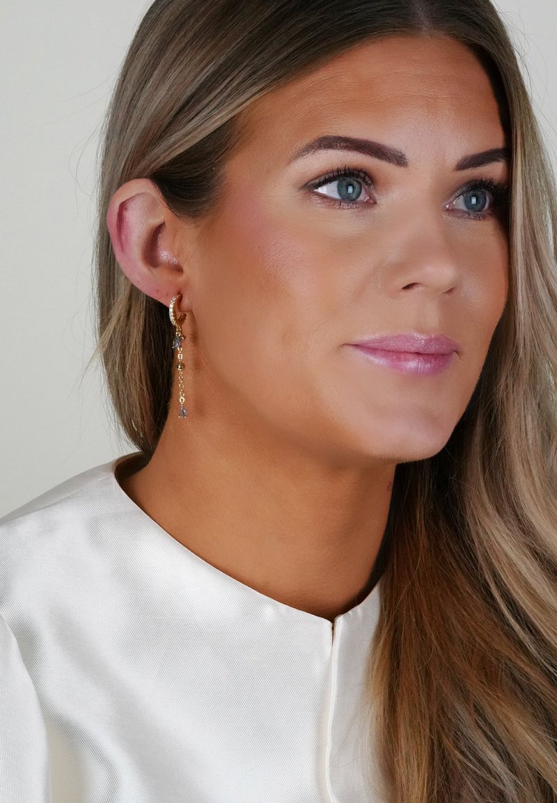 Guldtonade örhängen med hängande kedjor och blå ädelstenar. Bärs med en satinfärgad vit topp som framhäver accessoarens design.