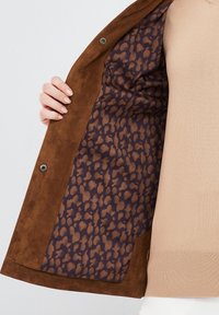 Bruine suède jas met een binnenkant van luipaardprint in tinten van donkerpaars en bruin. Heeft een knoopsluiting en een gevoerde textuur.