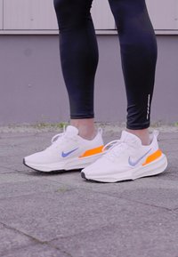 Scarpe da ginnastica bianche con accenti blu e dettagli arancioni. Il design presenta una parte superiore testurizzata e una suola ammortizzata per offrire supporto.