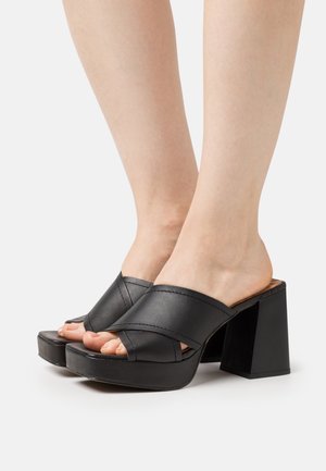 Mulas de plataforma negras con diseño de correas cruzadas, textura de cuero suave, tacón cuadrado y abierto que muestra los dedos pulidos.