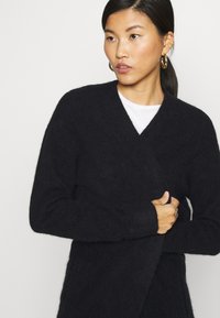Svart strikket cardigan med dyp V-hals, lange ermer og myk tekstur. Har en omslagsdesign og subtile sømdetaljer.