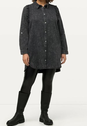 Mujer con camisa larga negra con botones y hombros con volantes, leggings de cuero negro y botas negras hasta la rodilla, posando frente a un fondo blanco.