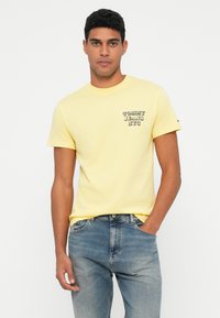 Geltona medvilninė marškinė su maža grafika kairėje krūtinės pusėje. Derinama su šviesiai mėlynomis džinsais. Paprastas apvalus kaklas ir trumpomis rankovėmis.