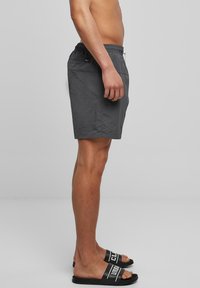 Grå badeshorts med elastisk midje og snor. Har en sidelomme og et lett, teksturert stoff. Sorte sandaler med hvite bokstaver.