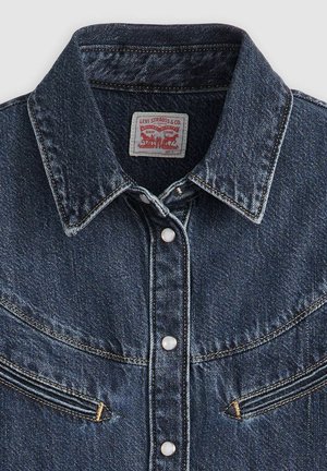 Dunkelblaue Denimjacke mit spitzem Kragen, zwei Brusttaschen und Knopfverschluss, der weiße Knöpfe und kontrastierende gelbe Nähte aufweist.